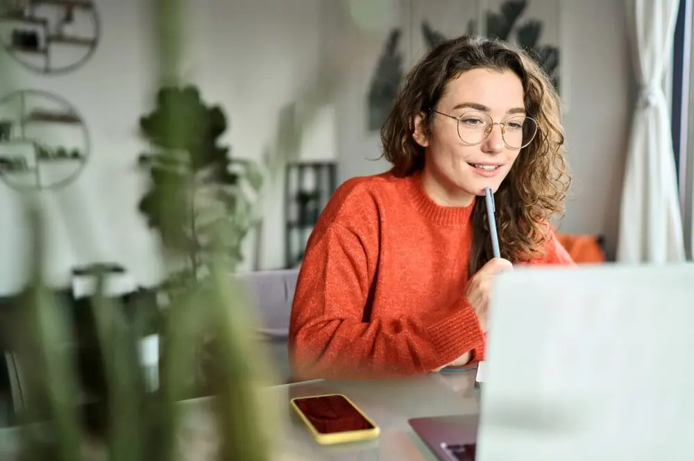 A woman with brown hair, wearing glasses and an orange sweater, presses a pen against the tip of her lower lip, and smiles calmly but excitedly while looking at a laptop screen.