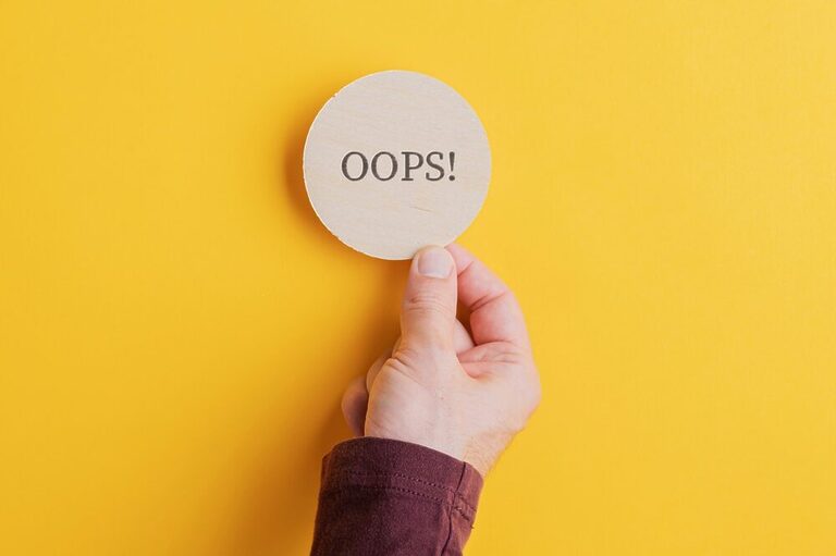 A hand in a yellow background holding a piece of paper saying "OOPS!"