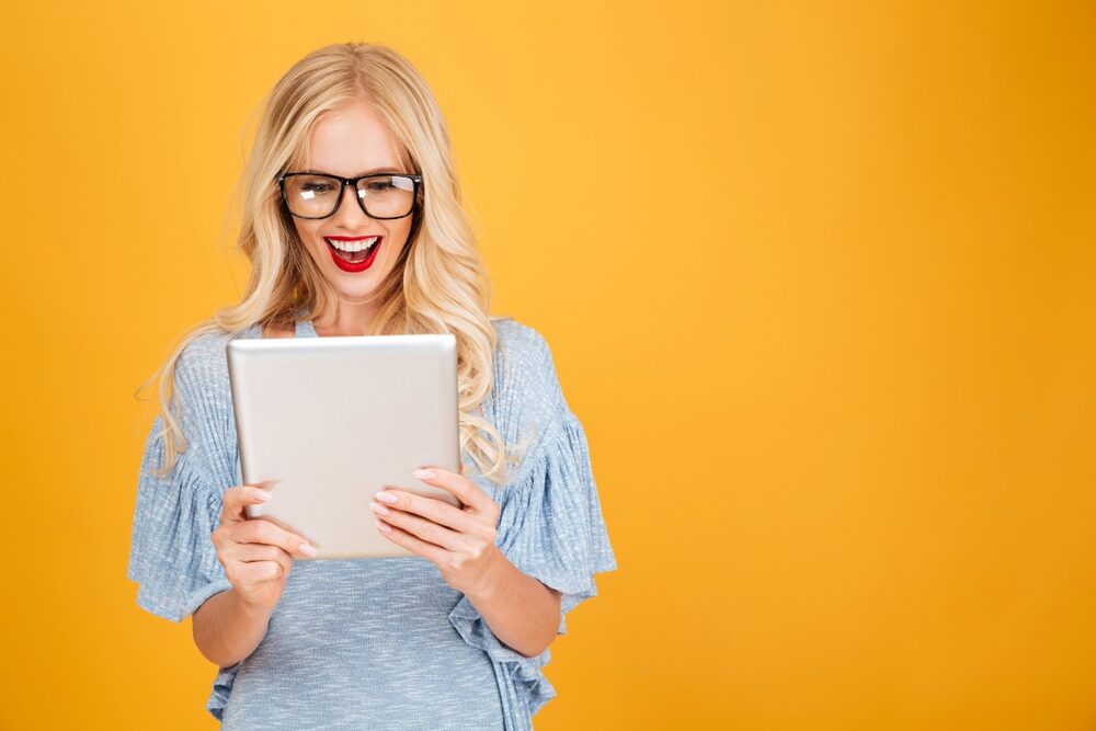 A blonde woman wearing a light blue blouse and glasses is holding a tablet and smiling excitedly as she looks at the screen. The woman is standing in front of a yellow wall, which contrasts with the light blue of her flowing blouse.