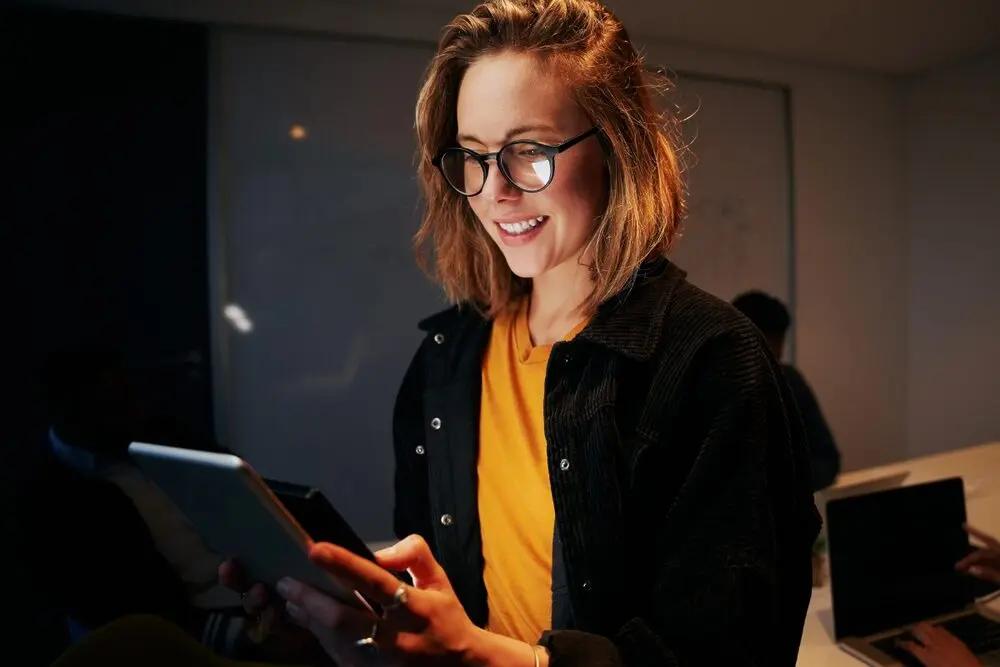 The photo shows a young blonde woman in a casual, modern corporate setting, wearing an orange shirt with a denim jacket over it, and glasses while checking something interesting on a tablet.