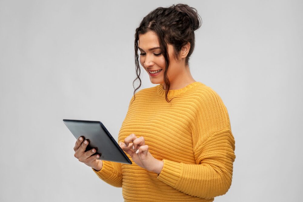 The photo shows a woman with curly hair, wearing a yellow sweater and focused on a tablet screen.