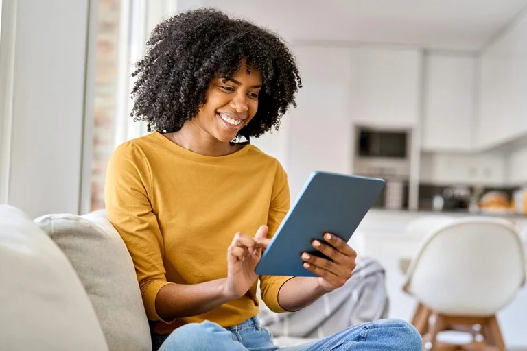 A woman, wearing a yellow blouse, sits on the sofa in her living room, opposite her kitchen, browsing on her tablet and smiling confidently.