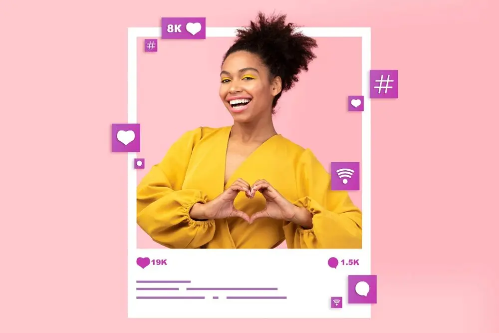 The colorful and fun image, reminiscent of an Instagram post, shows a woman with colorful clothes and makeup, contrasting with the light pink background, making the shape of a heart with both hands while smiling animatedly at the camera.