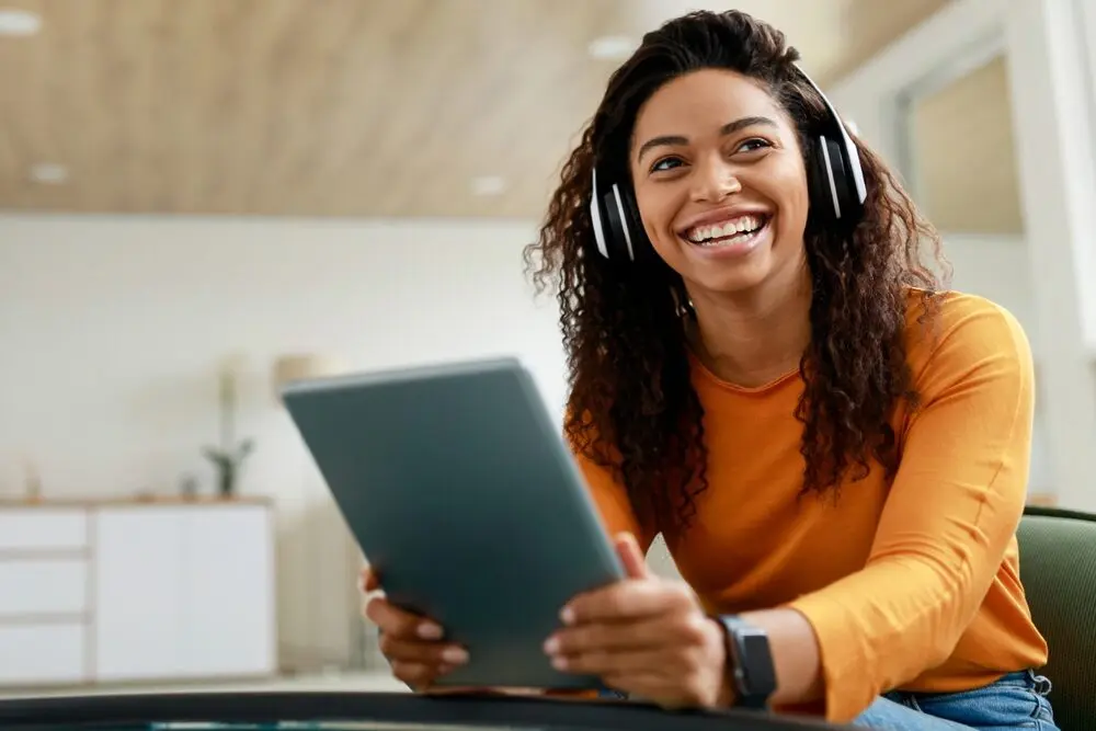 The image shows a black woman smiling animatedly while holding a tablet and wearing a long-sleeved orange blouse.