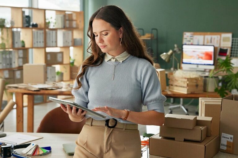 A woman wearing a gray sweater is checking something on her tablet inside an office with a storage area for packages and boxes for e-commerce.