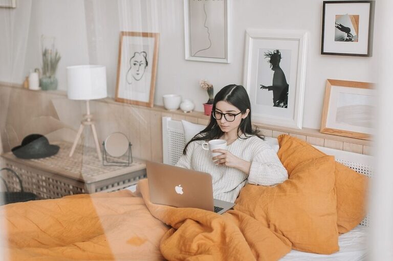 The image shows a brunette woman wearing glasses, sitting on a bed under the blankets, working on her laptop and drinking a cup of coffee.